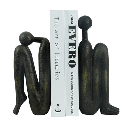 Pair of dark resin abstract human sculptures positioned on either side of two white books, with one figure leaning forward and the other standing upright.