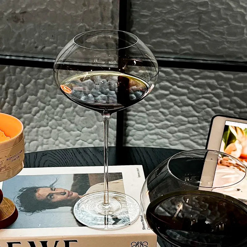  "Gravity" disc bowl crystal wine glass filled with a drink resting on a book on a table, distinctive flat disc bowl and elegant stem — CL House Drinkware collection