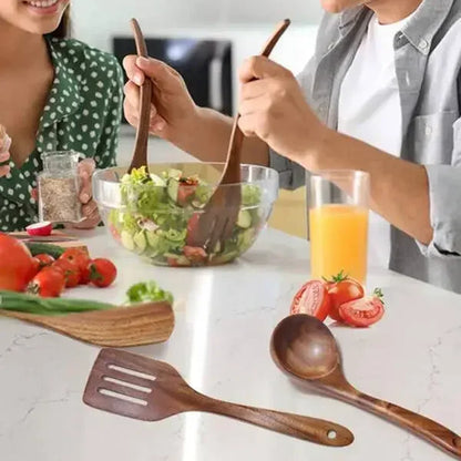 Couple tossing a fresh green salad together using teak wood serving spoons at a white marble kitchen counter — slotted spatula and ladle resting on the countertop alongside cherry tomatoes and orange juice — lifestyle shot of the Teak Grove Utensil Set by CL House