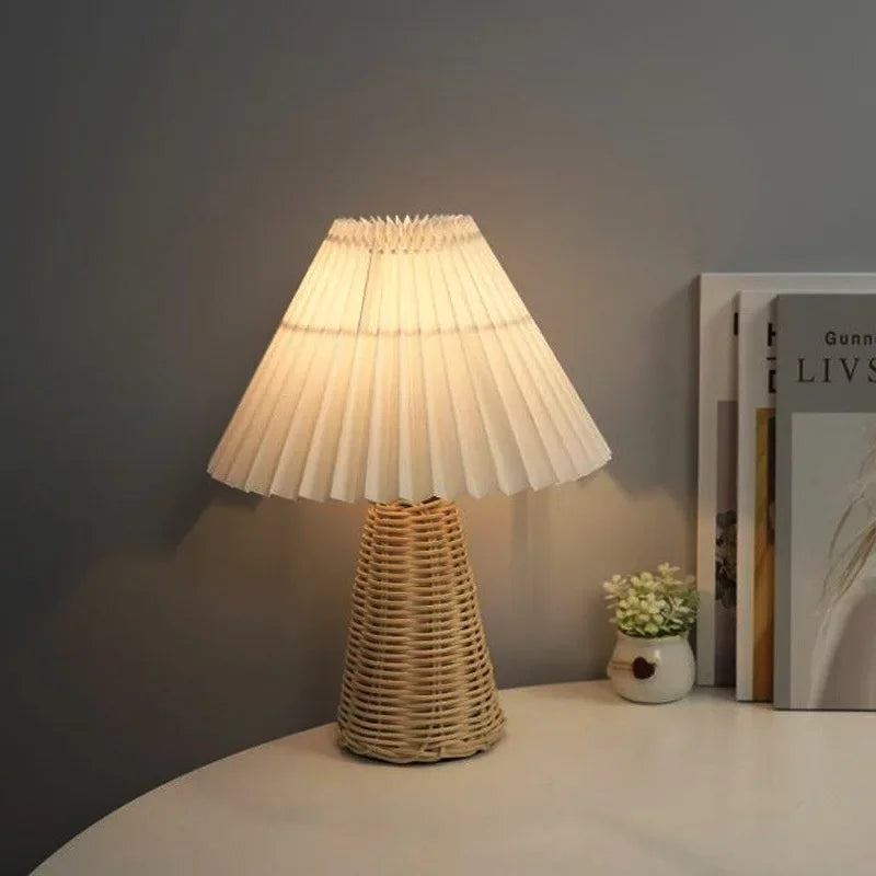  Rattan & Pleat table lamp with woven rattan cone base and wide off-white pleated shade casting warm light on a white desk, styled with a home decor magazine and a small ceramic vase with white flowers against a grey wall