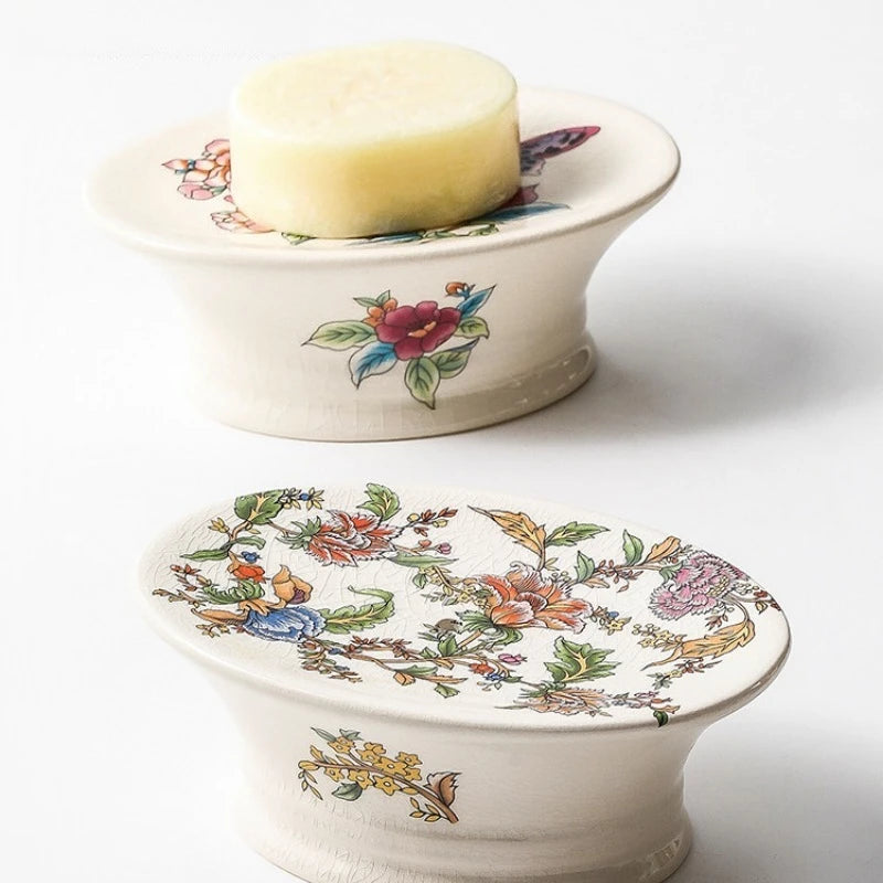 Two oval pedestal floral ceramic soap dishes — Butterfly Garden variant with bar of soap on top, Chinoiserie Garden variant below showing full botanical print, on white background