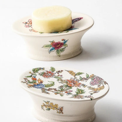 Two oval pedestal floral ceramic soap dishes — Butterfly Garden variant with bar of soap on top, Chinoiserie Garden variant below showing full botanical print, on white background