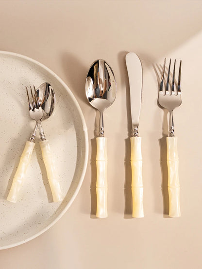 Jade Stem" Ivory Cream cutlery set — 5 pieces styled beside a speckled ceramic plate on a warm beige surface, showing dinner fork, knife, spoons and dessert fork — lifestyle shot