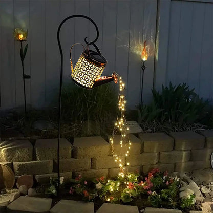 Starfall Kettle" solar fairy light watering can, Lattice Copper finish, mounted on shepherd's hook pole in a night garden, warm LED fairy lights cascading from spout onto a stone flower bed, atmospheric outdoor lighting setting
