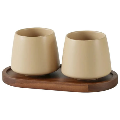 Two cone-shaped matte oat white ceramic cups on oval acacia wood tray, warm beige finish, bathroom toothbrush holder set, white background