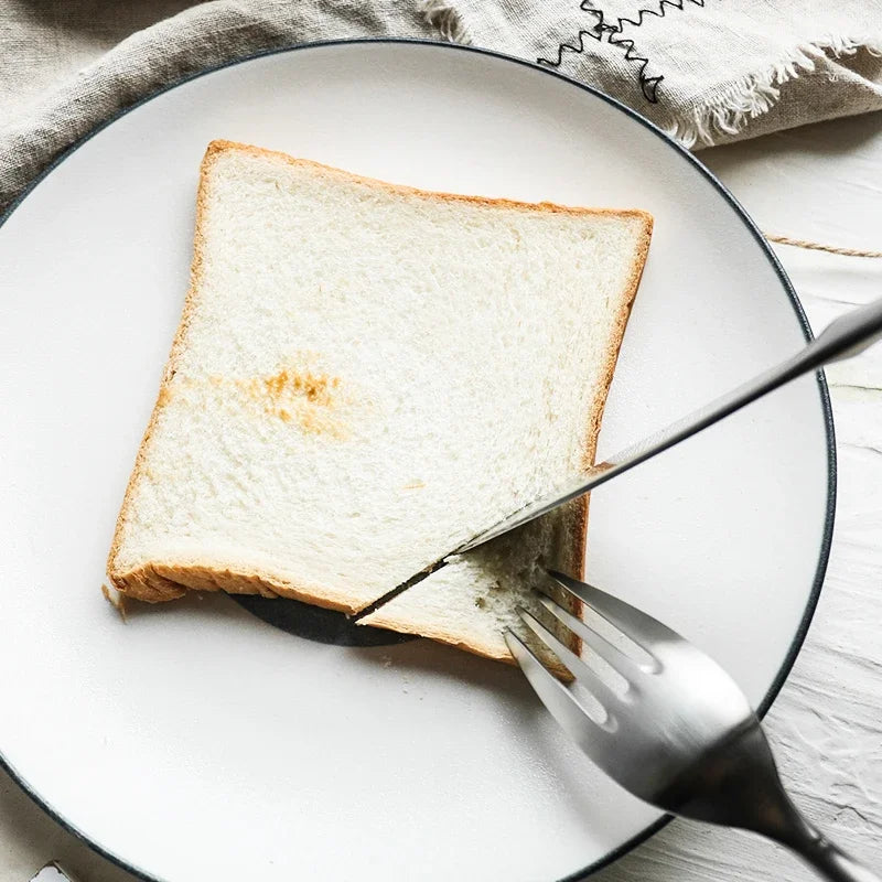  Stainless steel knife and fork with natural wood handles cutting bread on a white ceramic plate