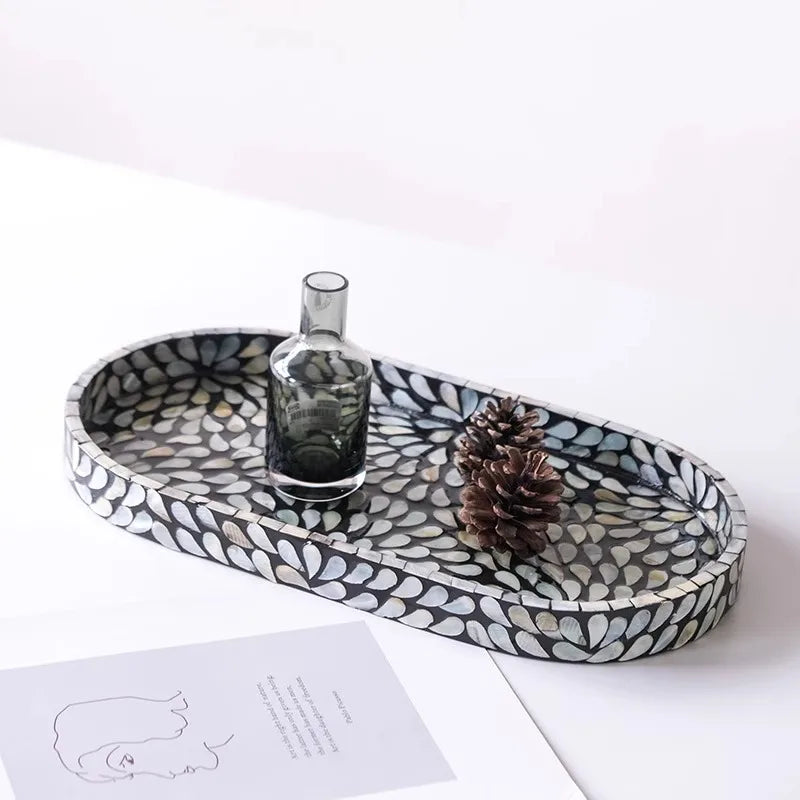 Midnight mother-of-pearl oval tray styled with smoky glass vase and pine cone on white surface, art magazine beside, iridescent shell mosaic floral pattern detail — luxury home decor