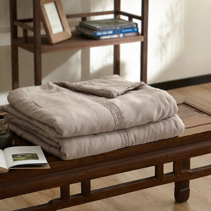 Warm coffee quilted linen cotton summer throw blanket, neatly folded on a dark wood bench in a japandi-style interior with wooden shelving and open book