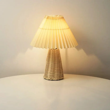  Rattan & Pleat table lamp with woven rattan base and layered off-white pleated shade, glowing warm light on a white surface against a neutral wall
