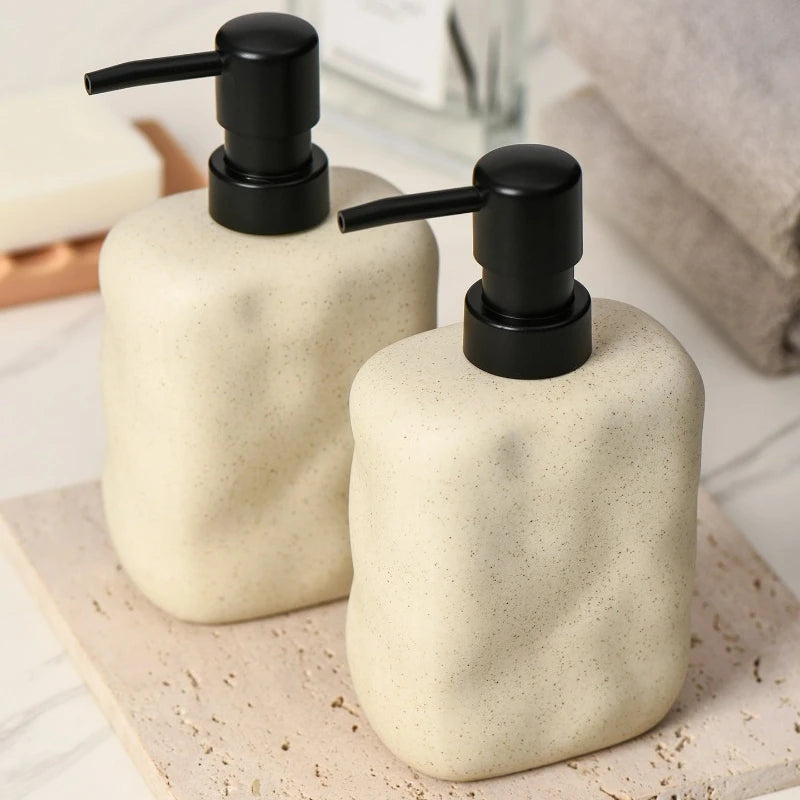 Two Sand Form ceramic soap dispensers with matte black pumps on travertine tray, styled bathroom vignette with white towel and marble background