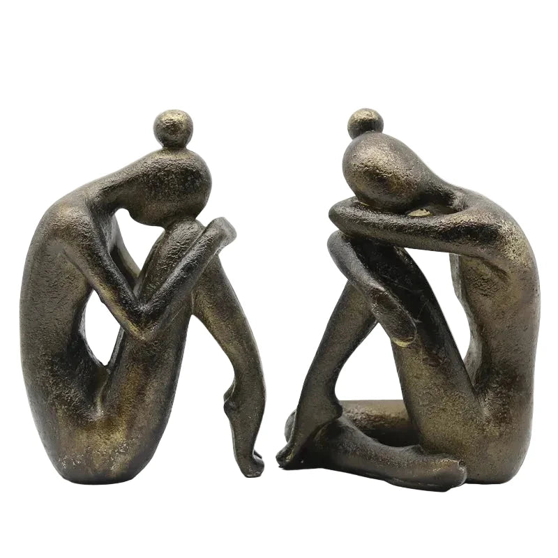 Pair of dark resin abstract seated figures displayed indoors in soft natural light, showing their different sculptural poses and textured finish.