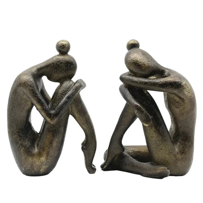 Pair of dark resin abstract seated figures displayed indoors in soft natural light, showing their different sculptural poses and textured finish.
