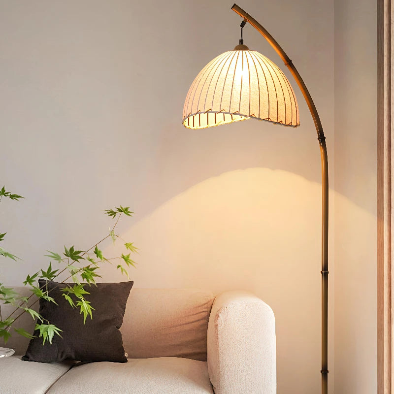 Arc & Pleat canvas floor lamp casting warm light over a cream sofa and dark cushion, with a Japanese maple plant beside it in a minimalist living room setting
