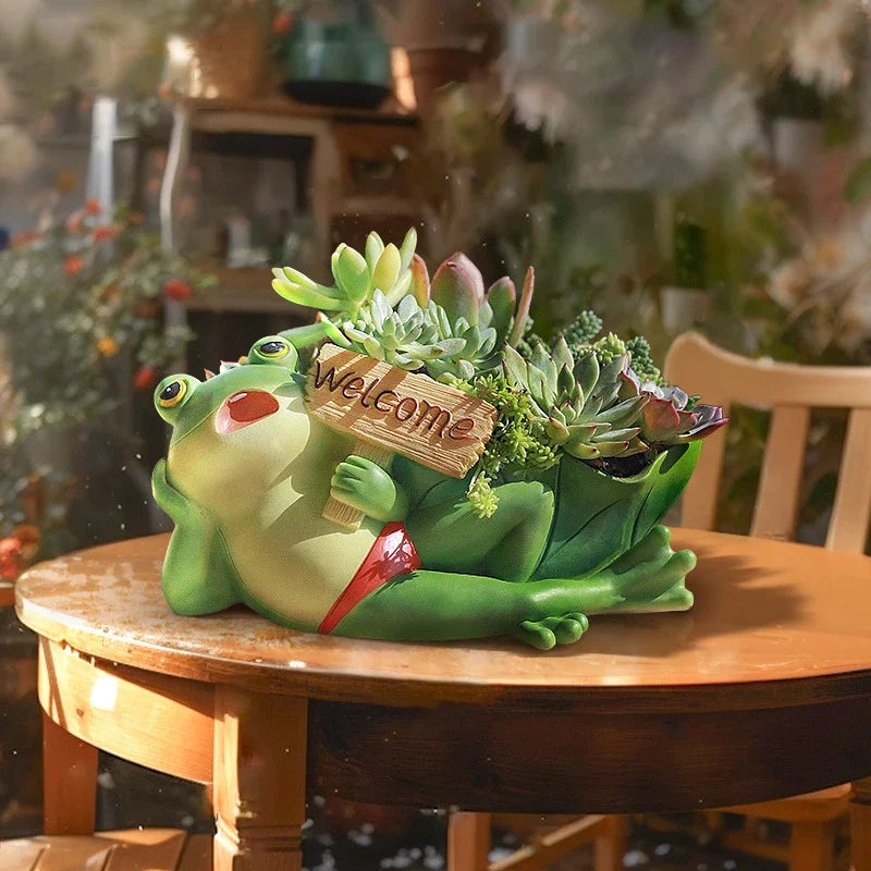 Frog Tale" resin frog planter — Welcome Lounger styled with mixed succulents on a wooden garden table in a greenhouse setting