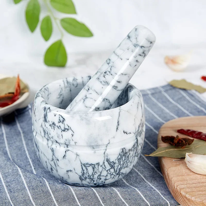 White marble mortar and pestle with dark grey veining on blue striped linen cloth — garlic cloves, dried red chillies, bay leaf and star anise scattered around, fresh green leaves in soft-focus background — lifestyle hero shot of the White Marble variant of the Stone Grind Mortar & Pestle Set by CL House