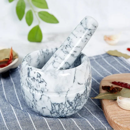 White marble mortar and pestle with dark grey veining on blue striped linen cloth — garlic cloves, dried red chillies, bay leaf and star anise scattered around, fresh green leaves in soft-focus background — lifestyle hero shot of the White Marble variant of the Stone Grind Mortar & Pestle Set by CL House