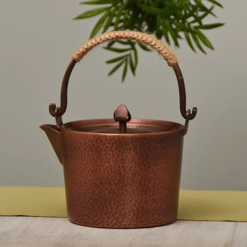 Copper Ritual handmade pure copper teapot kettle in Antique Copper finish styled on a linen table runner with a bamboo plant in the background, showing the deep hammer-textured patina body and jute-wrapped arched handle