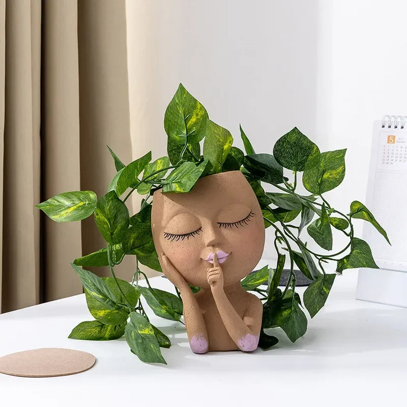"Hush Muse" sculptural face resin planter in warm terracotta with closed eyes, shh gesture and lilac nail detail, displayed with cascading golden pothos plant on white desk