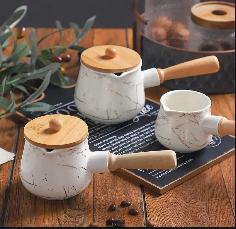 Three white marble ceramic milk jugs in all three sizes — small with bamboo lid, medium with bamboo lid, and large without lid — displayed on a dark slate board over a rustic wooden table, olive branch and coffee beans in the background — full size range of the Marble Pour Ceramic Milk Jug by CL House