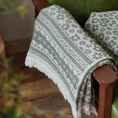Sage Bloom 100% cotton jacquard woven throw blanket with floral pattern and white fringe trim draped over a green leather armchair in a styled living room