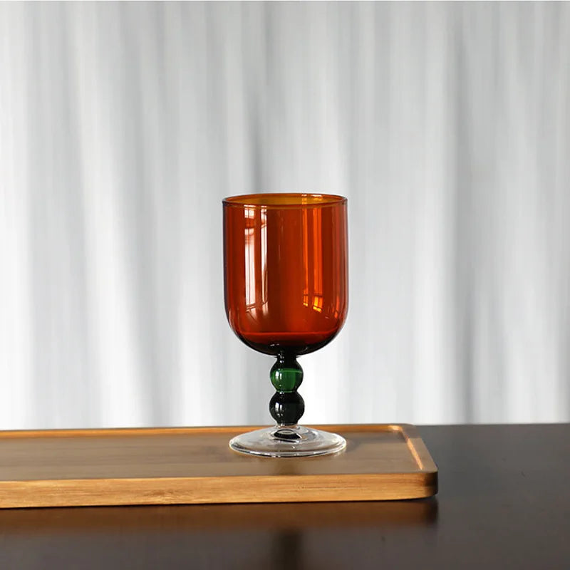 "Chromatic Orb" glass goblet in deep amber with two-tone green and black bead stem, on a wooden tray against a white curtain background