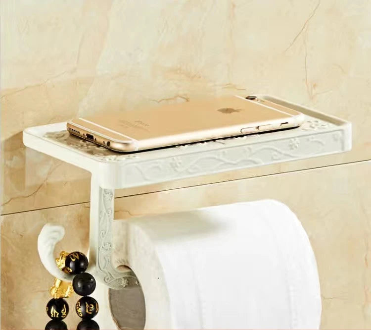 Wall-mounted ivory white toilet paper holder with engraved arabesque pattern and phone shelf, smartphone resting on top, marble wall background