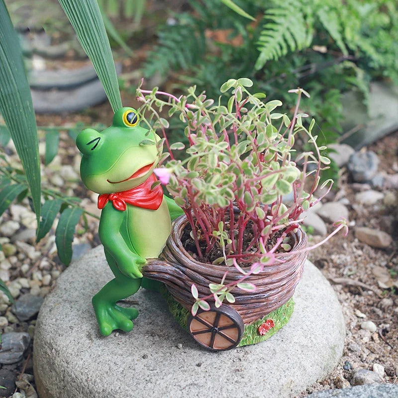 "Frog Tale" resin frog planter — Basket Hauler styled with trailing succulents resting on a garden stone among pebbles and ferns