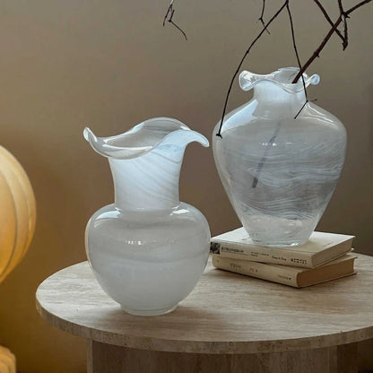 "Petal Wave" Milk Swirl and Cloud Drift handblown glass vases side by side on round wooden table — Cloud Drift holding bare winter branches, warm ambient light