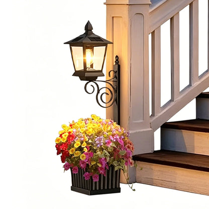 Solar garden post lantern in matte black glowing with warm white light, wall-mounted beside a staircase with a planter box overflowing with colourful mixed flowers