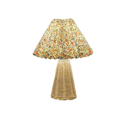  Rattan & Pleat table lamp with woven rattan cone base and pleated floral shade in red, orange and green botanical print, warm light on a white surface against a taupe wall
