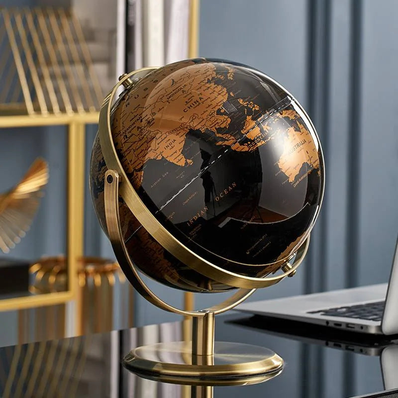 Decorative world globe with black and gold vintage map on gold-plated stand, styled on a glass desk next to a laptop in a modern home office with blue wall and gold shelving