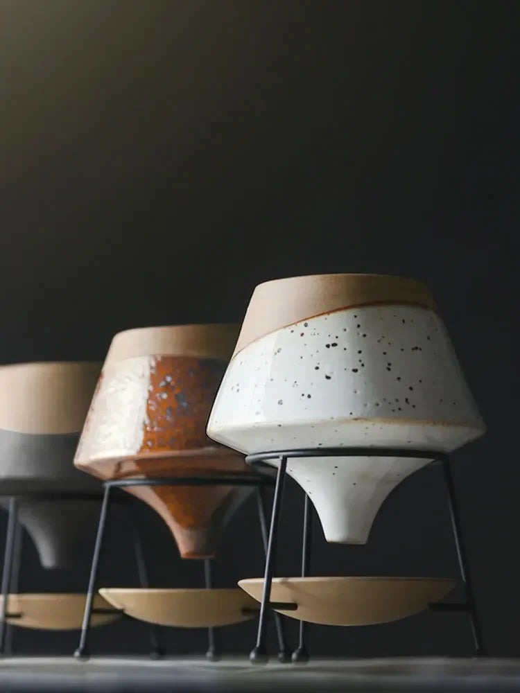  All three Ember Cone ceramic plant pots on black iron tripod stands displayed together — slate and sand, amber glaze and speckled white colourways — dark background, dramatic studio lighting