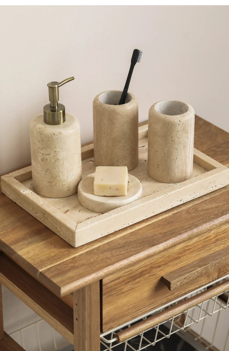 Imagem 1: Natural travertine bathroom set on a wooden bedside table — soap dispenser with brass pump, toothbrush cup, mouthwash cup, round soap dish and rectangular tray in warm beige stone — wabi-sabi bathroom styling