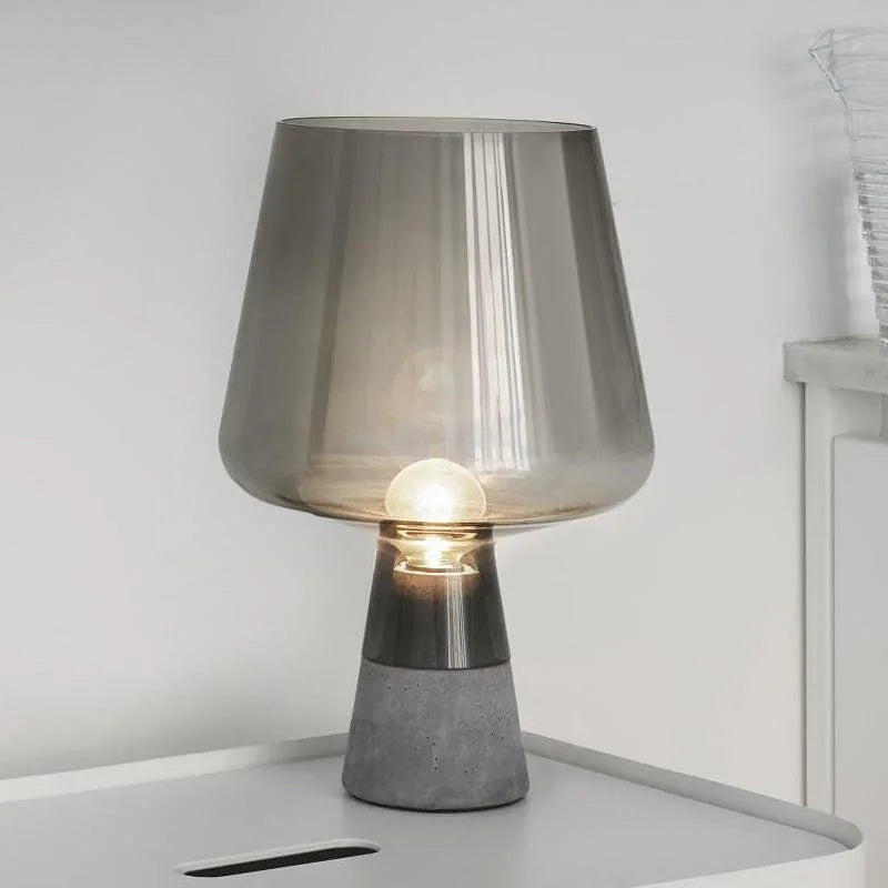"Smoke & Cement" glass table lamp with smoke grey shade on a white desk, warm bulb glowing against a light grey wall