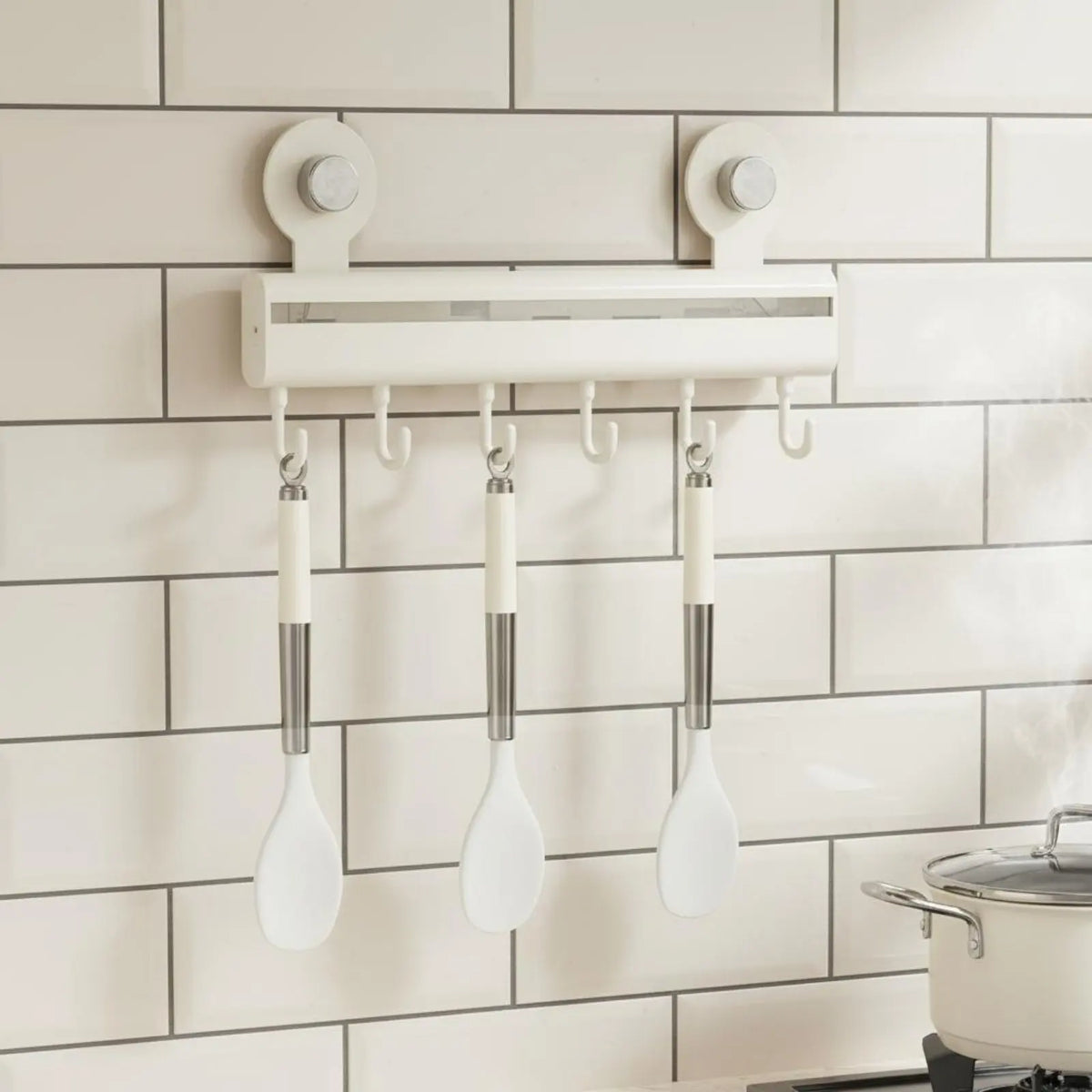 Cream adhesive wall-mounted kitchen hook rail with seven sliding hooks holding white silicone spoons on white subway tiles with a stainless steel pot on the hob below, CL House & Home
