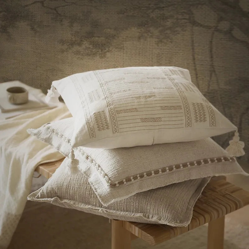 Ivory tassel jacquard square cushion cover stacked with a natural linen cushion cover on a wooden stool, styled with a linen throw and ceramic mug against a rustic stone wall — boho wabi-sabi home decor.