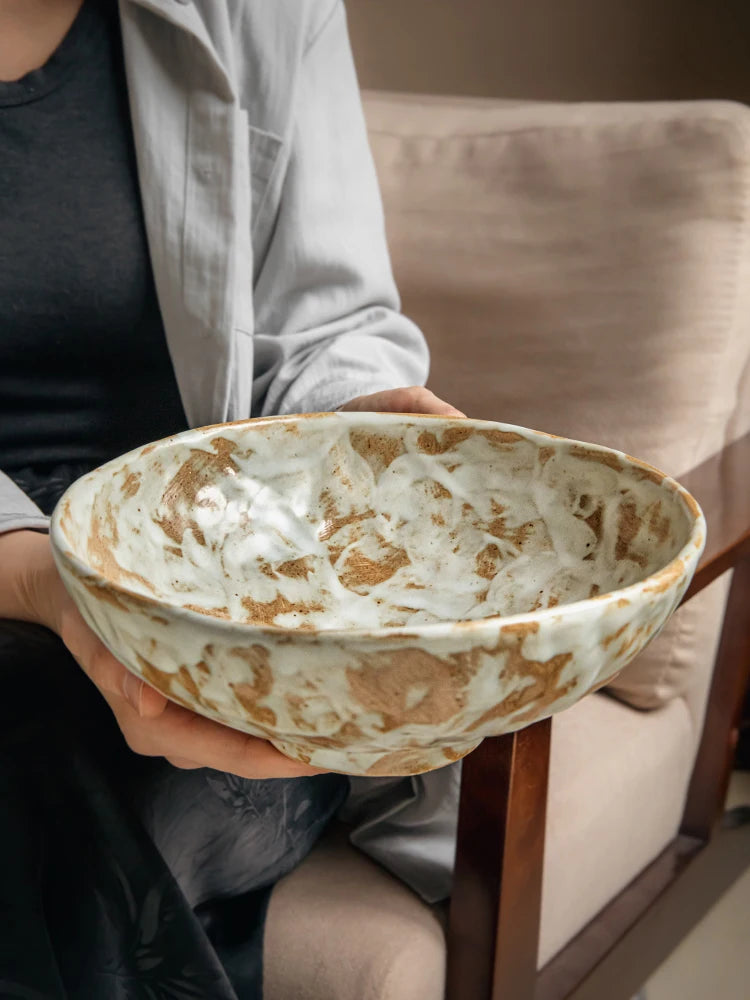 Large rustic stoneware serving bowl with amber brushstroke glaze, exterior view showing cream and amber reactive glaze texture, held by person seated on beige sofa