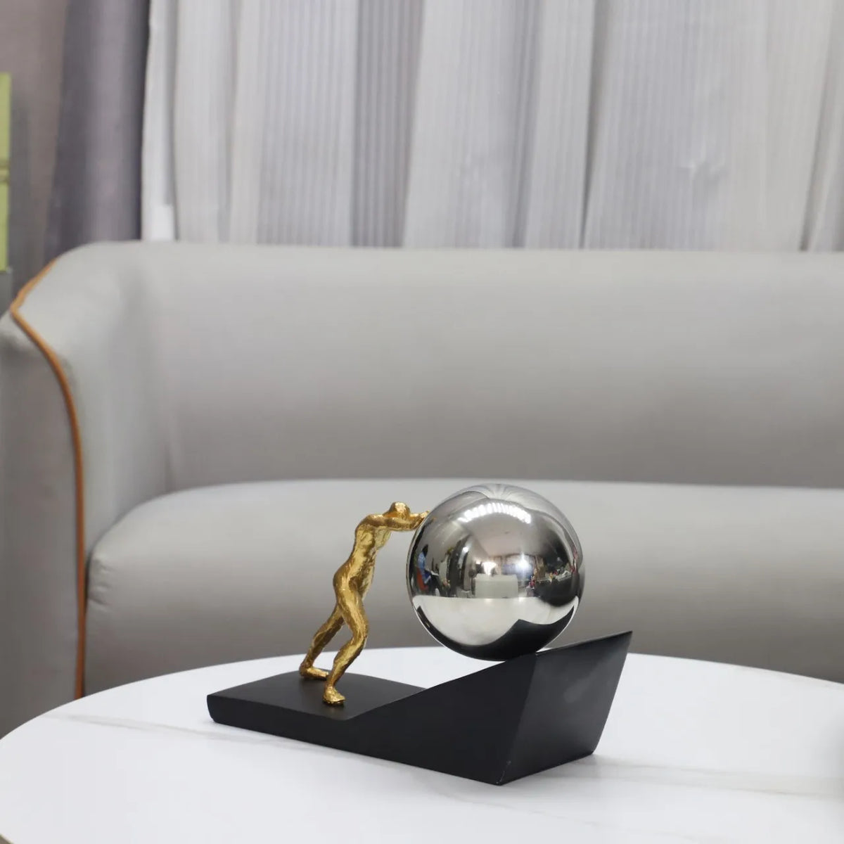 Silver sphere version of The Ascent metal Sisyphus sculpture displayed on a white coffee table in front of a cream sofa.⠋