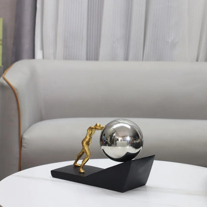 Silver sphere version of The Ascent metal Sisyphus sculpture displayed on a white coffee table in front of a cream sofa.⠋