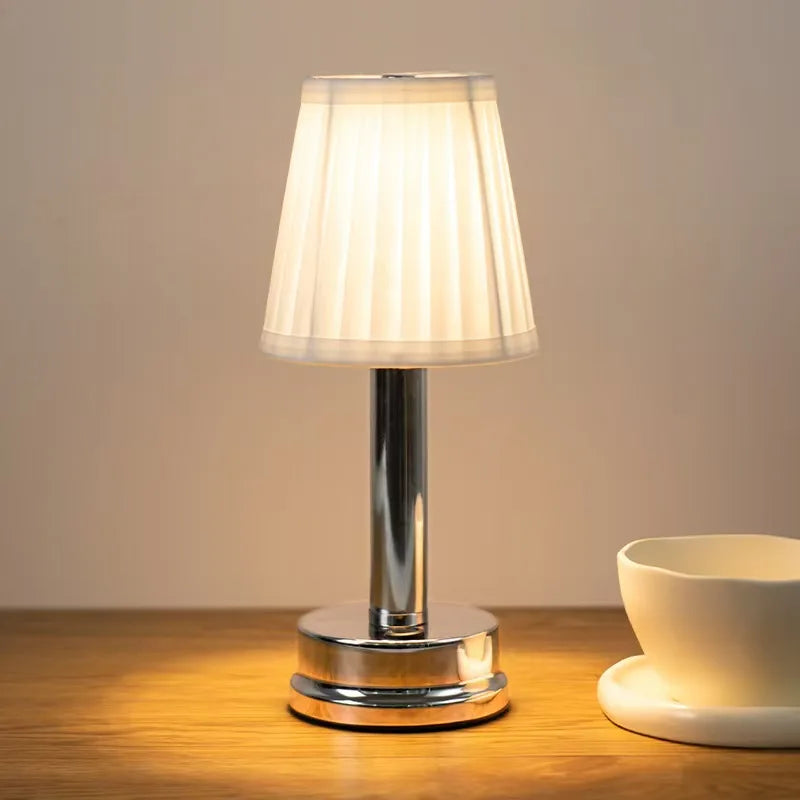  Silver rechargeable touch table lamp with pleated fabric shade glowing warm white on a wooden surface, against a neutral beige background