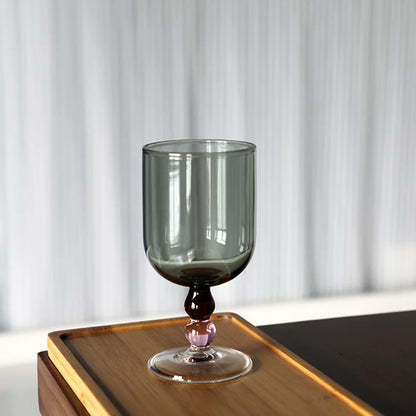 "Chromatic Orb" glass goblet in smoke grey with two-tone wine and pink bead stem, on a wooden tray against a white curtain background