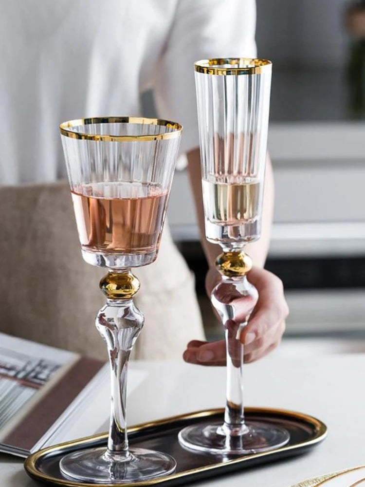 Hand holding a "Golden Hour" crystal wine glass filled with red wine alongside a champagne flute, gold filigree rim detail — CL House Drinkware collection