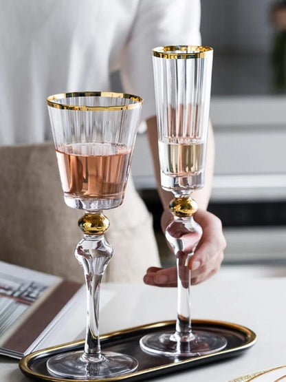Hand holding a "Golden Hour" crystal wine glass filled with red wine alongside a champagne flute, gold filigree rim detail — CL House Drinkware collection