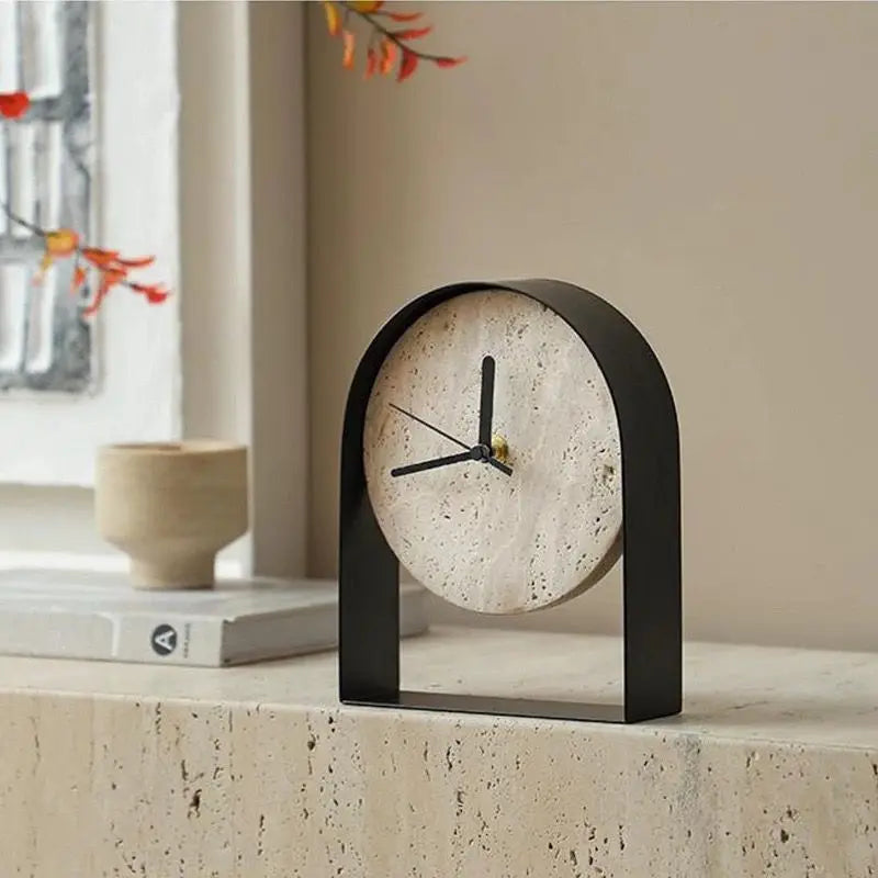 Black arched travertine table clock with black hands on a travertine ledge, beside a stone bowl and a book by a window