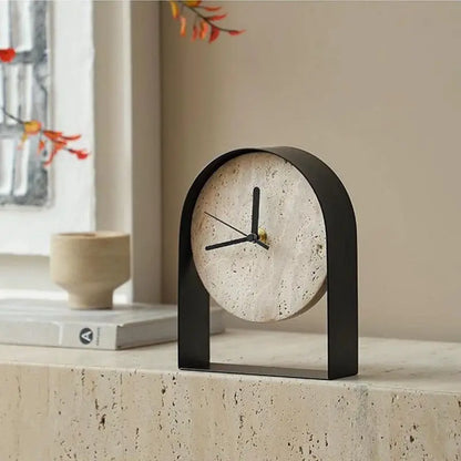 Black arched travertine table clock with black hands on a travertine ledge, beside a stone bowl and a book by a window