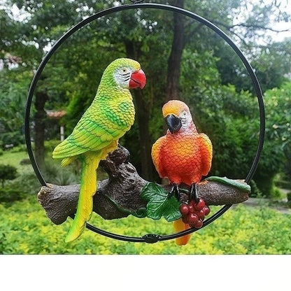"Perch Duo" resin parrot hanging ring ornament in Emerald & Amber — hand-painted green and orange parrots perched on a driftwood branch with berry detail, garden background