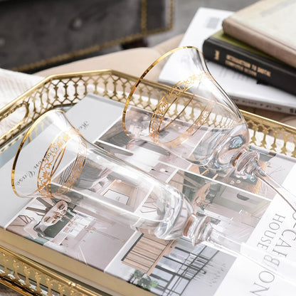 "Golden Filigree" crystal wine glass and champagne flute on brass tray in elegant living room setting with books and greenery
