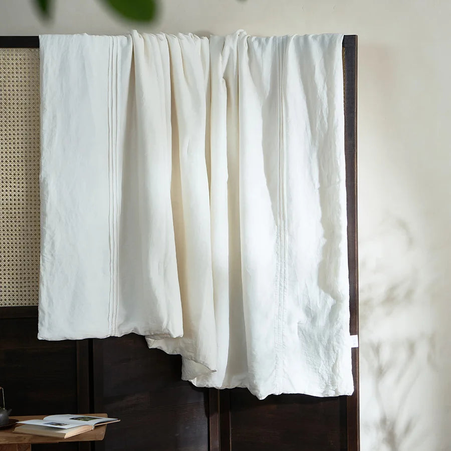  Ivory white quilted linen cotton summer throw blanket, neatly folded on a dark wood bench in a japandi-style interior with wooden shelving and open book