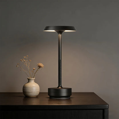 Black disc-shade rechargeable table lamp illuminated on a dark wooden surface, styled with a small ceramic vase and dried flowers — "Disc & Stem" by CL House
