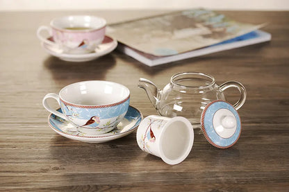 Blue "Happy Bird" tea for one set disassembled on a wooden table — porcelain cup with saucer, glass teapot, porcelain infuser and lid displayed separately, with pink cup and magazine softly blurred in the background.